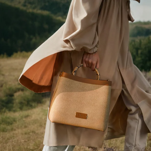 Person in a flowing beige dress carries a woven tan handbag through a sunlit meadow, with wooded hills behind, creating a serene mood.