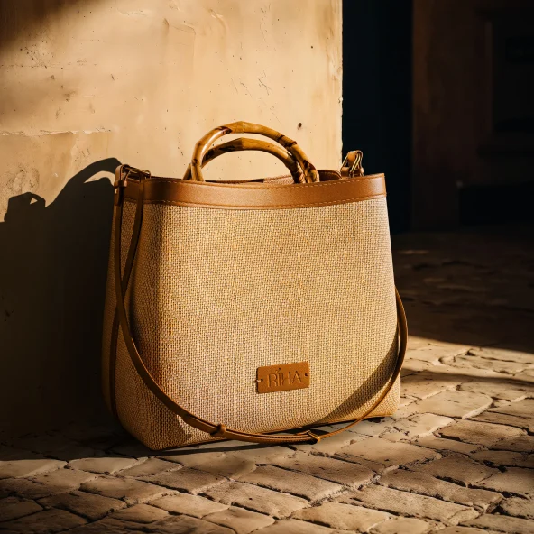 Tan woven leather tote with bamboo handles rests in warm sunlight on a rustic brick floor beside a weathered wall, casting shadows.