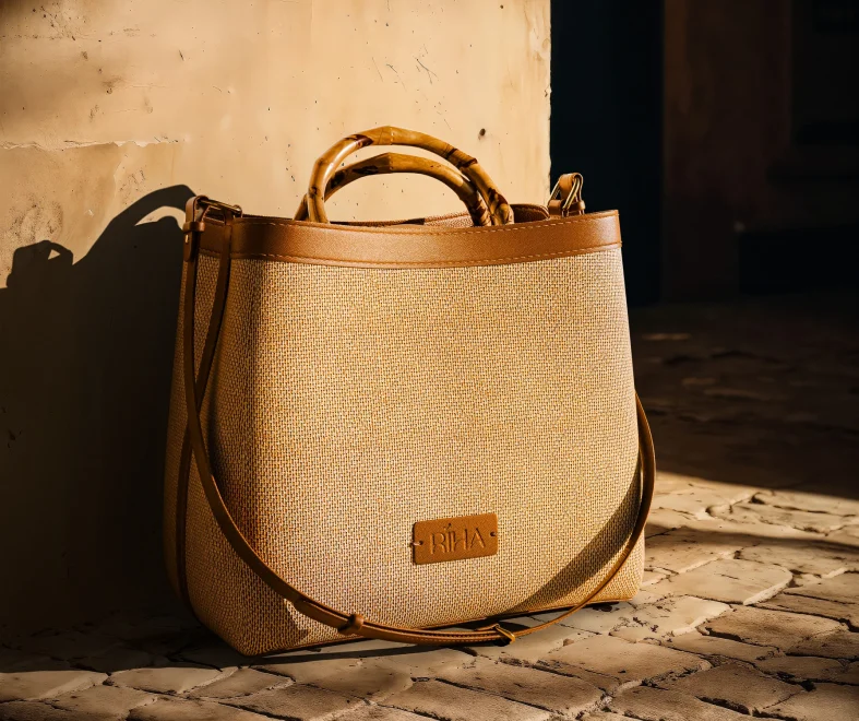 Tan woven leather tote with bamboo handles rests in warm sunlight on a rustic brick floor beside a weathered wall, casting shadows.