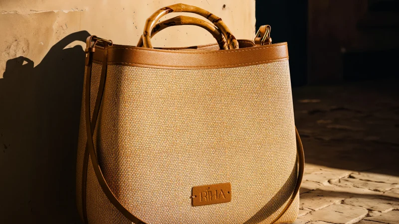 Tan woven leather tote with bamboo handles rests in warm sunlight on a rustic brick floor beside a weathered wall, casting shadows.