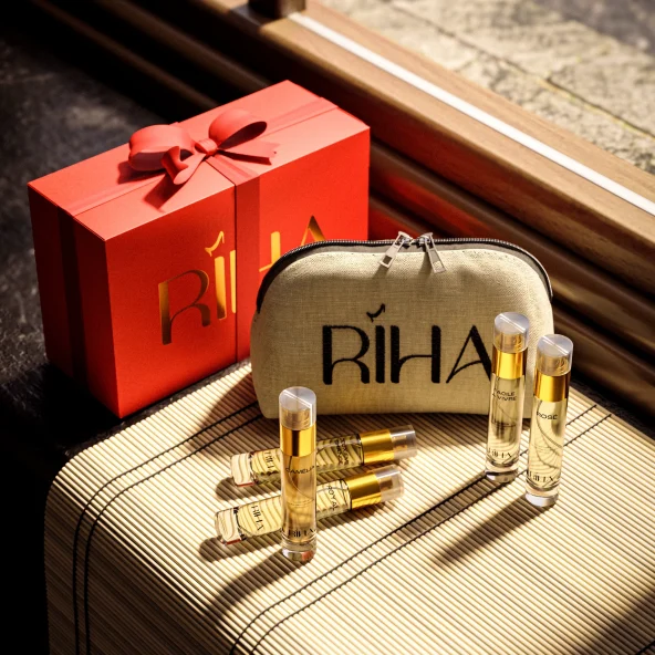 Luxury perfume gift set with a red ribboned box, beige RİHA pouch, and gold-capped fragrance vials on a sunlit surface; warm, elegant tone.