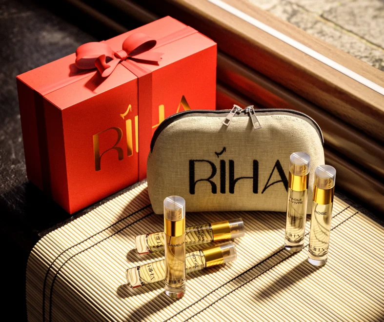 Luxury perfume gift set with a red ribboned box, beige RİHA pouch, and gold-capped fragrance vials on a sunlit surface; warm, elegant tone.