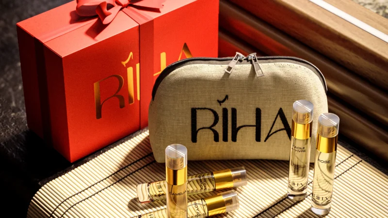 Luxury perfume gift set with a red ribboned box, beige RİHA pouch, and gold-capped fragrance vials on a sunlit surface; warm, elegant tone.