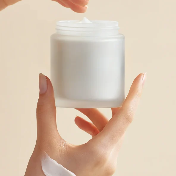 Hands hold an open white skincare cream jar, with a dab of lotion on the wrist, conveying clean, gentle self-care against a soft beige background.