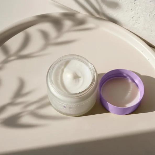 Open jar of white skincare cream beside a purple lid on a sunlit surface, with soft plant shadows creating a calm, serene mood.