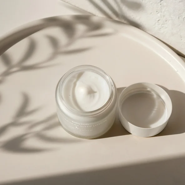 Open glass jar of white cream beside its lid on a sunlit surface, with soft leaf shadows creating a warm, minimalist skincare scene.