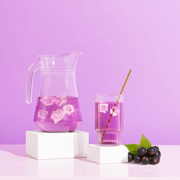 Clear pitcher and glass hold a pink iced drink with straws, beside black grapes and white blocks against a playful purple-pink backdrop.