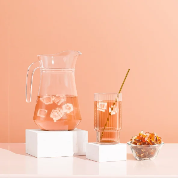 Pastel peach still life with a pitcher of iced orange drink, matching glass and straw, plus colorful gummy candies; playful and sunny.