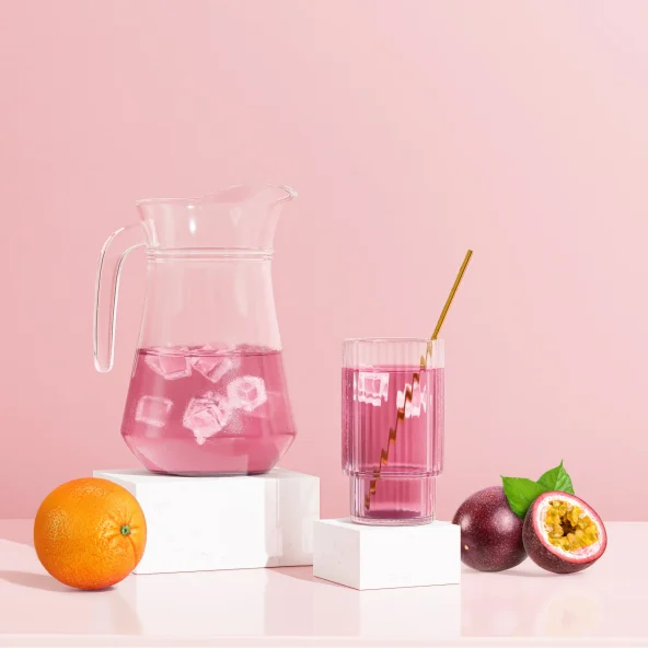 A glass and pitcher of pink juice with ice sit beside an orange and halved passion fruit on a pink surface, with a pink background.