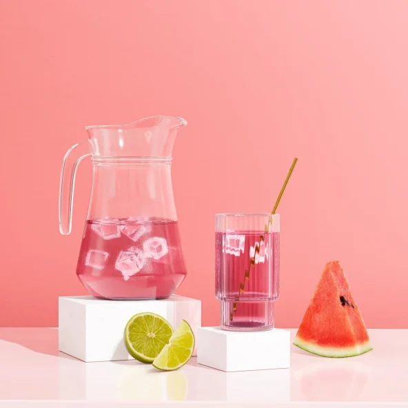 A glass pitcher and a glass of pink drink with ice, a straw, lime halves, and a watermelon slice on white blocks against a pink background.