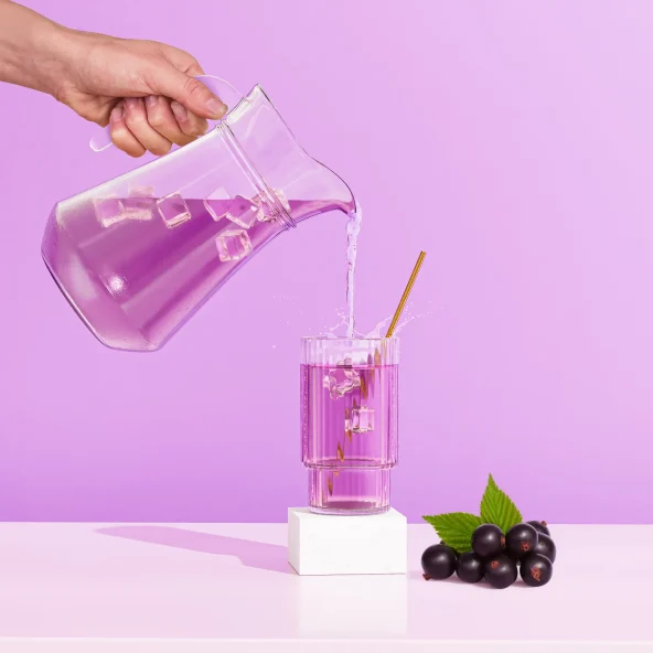 Hand pours water from a glass pitcher into a pink drink with ice and straw; blackberries sit on a white pedestal against a purple backdrop.