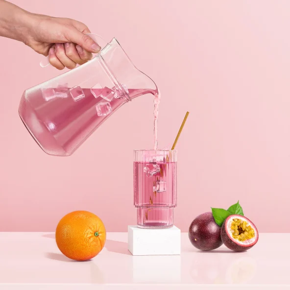 A hand pours pink juice from a glass pitcher into a tall glass with ice and a straw, with an orange and passion fruit on a white surface against a pink background.