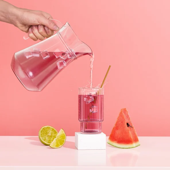 A hand pours pink iced drink from a glass pitcher into a straw-filled glass, with an orange and passion fruits beside it.